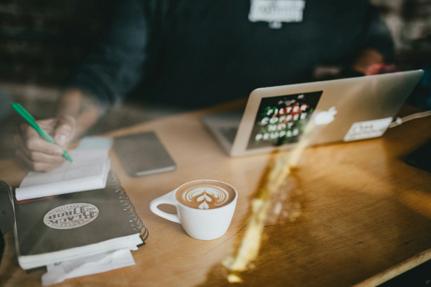 Persona che scrive su un taccuino a un tavolo di legno con laptop e caffè.